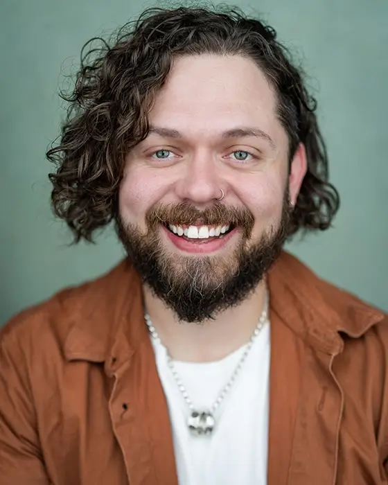 Keith Higinbotham in a brown shirt and white t-shirt with jaw-length curly hair, beard and moustache. His nose and ear is pierced.