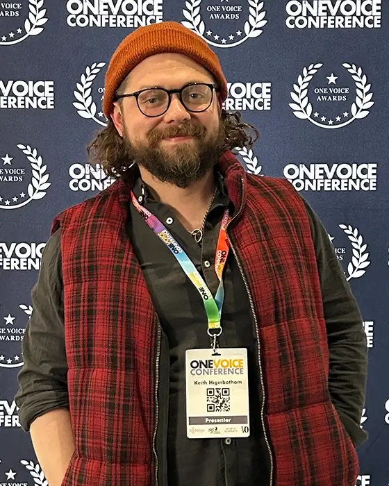 Keith Higinbotham with a One Voice Conference presenter lanyard on, stood in front of a logo step and repeat banner.