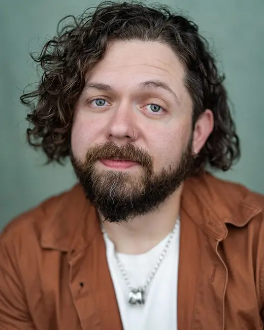 Keith Higinbotham in a brown shirt and white t-shirt with jaw-length curly hair, beard and moustache. His nose and ear is pierced.