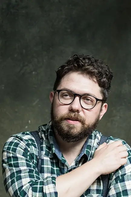 Keith Higinbotham looking away from the camera wearing a plaid shirt, braces with his right hand on his left shoulder.